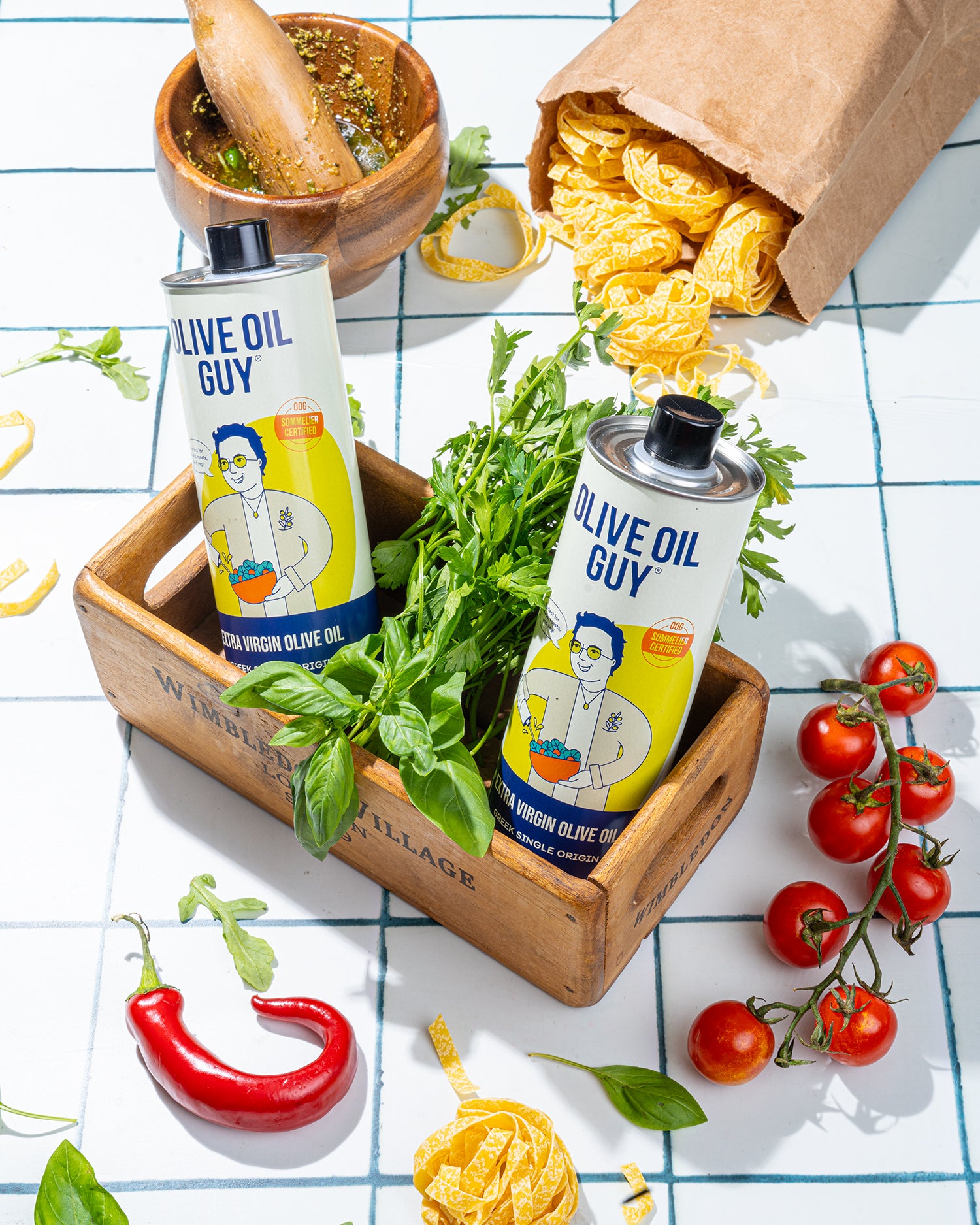 Two bottles of Olive Oil Guy on a wooden tray with herbs, tomatoes, and pasta.