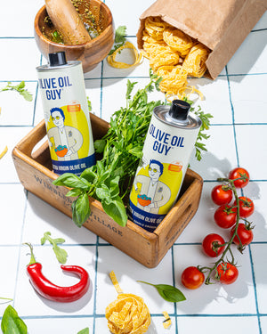 Two bottles of Olive Oil Guy on a table with fresh herbs, tomatoes, and pasta.
