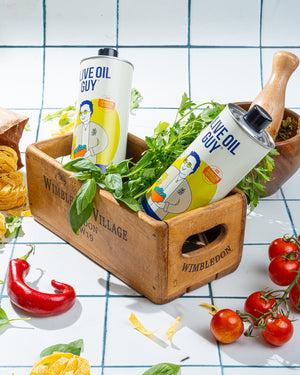 Two bottles of 'Olive Oil Guy' on a wooden tray with fresh herbs and vegetables on a checkered tablecloth.