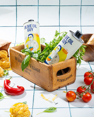 Two cans of 'Olive Oil Guy' on a wooden crate with vegetables and snacks on a checkered tablecloth.