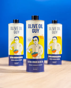 Three bottles of Olive Oil Guy olive oil on a wooden surface with a blue background