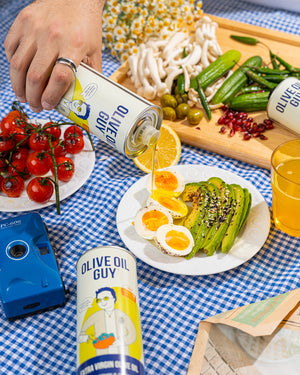 Olive Oil Guy can being poured over a salad on a table with vegetables and a camera.