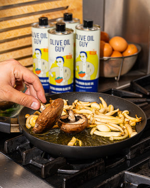 Mazen Assaf is cooking mushrooms and fries in a pan with 'Olive Oil Guy' olive oil bottles in the background.