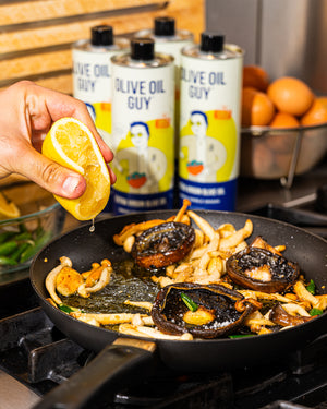 Person squeezing lemon over food in a pan with 'Olive Oil Guy' olive oil cans in the background.