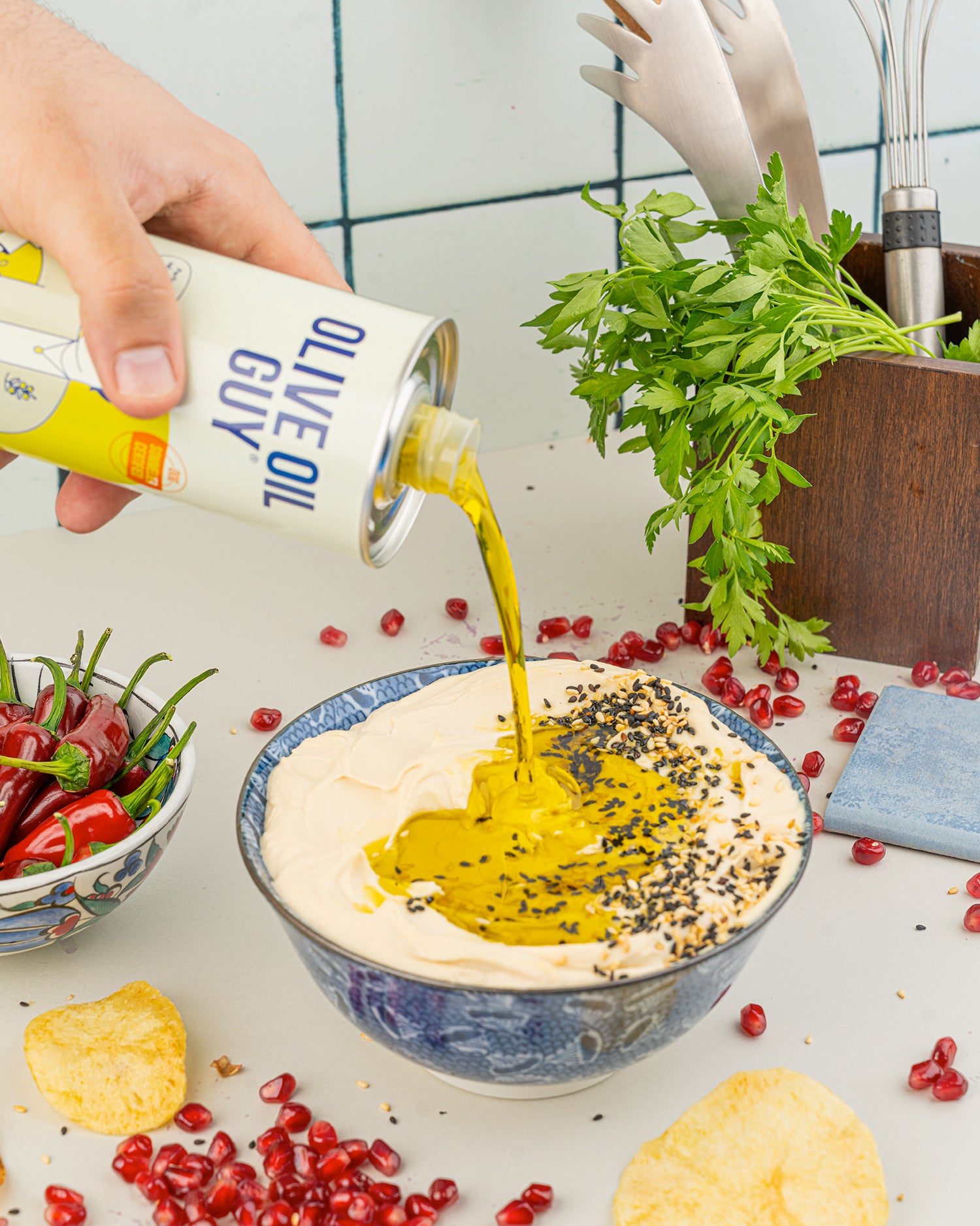 Olive oil being poured into a bowl of hummus with pomegranate seeds and crackers on a kitchen counter.