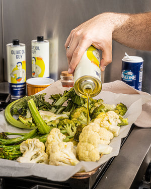 Mazen Assaf is pouring olive oil over roasted vegetables on a baking sheet with 'Olive Oil Guy' bottles in the background.