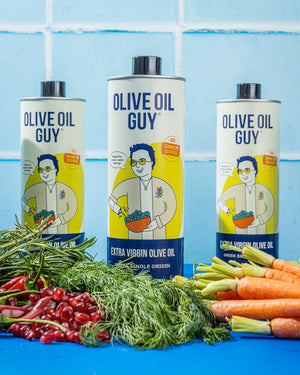 Three bottles of Olive Oil Guy olive oil with vegetables on a blue background